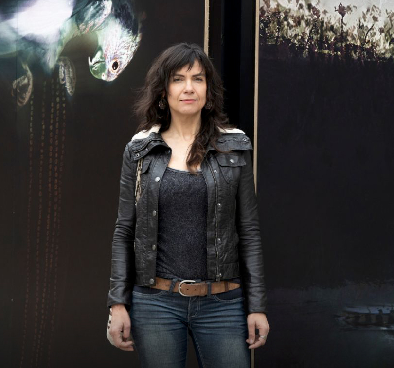 Judith Nangala Crispin - A woman standing outside (at the Australian National University), against a dark background, she has dark shoulder length hair, and is wearing a black leather jacket, and jeans, and looks strong. The background shows parts of her lumen artwork, we see  the head and beak of a bird in the top left hand corner.