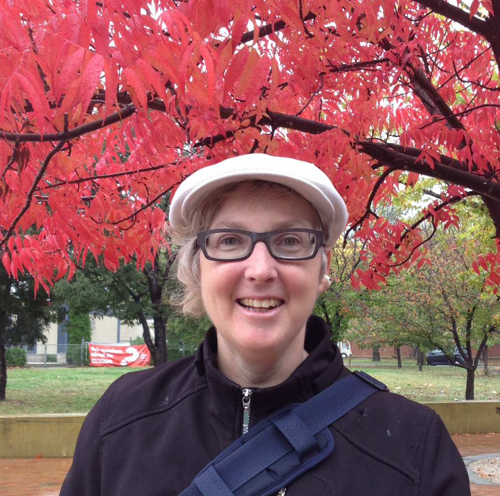 Sarah Rice - A smiling woman with short grey curly hair a white peaked cap and glasses, and in the background is vivid red Autumn maple trees.