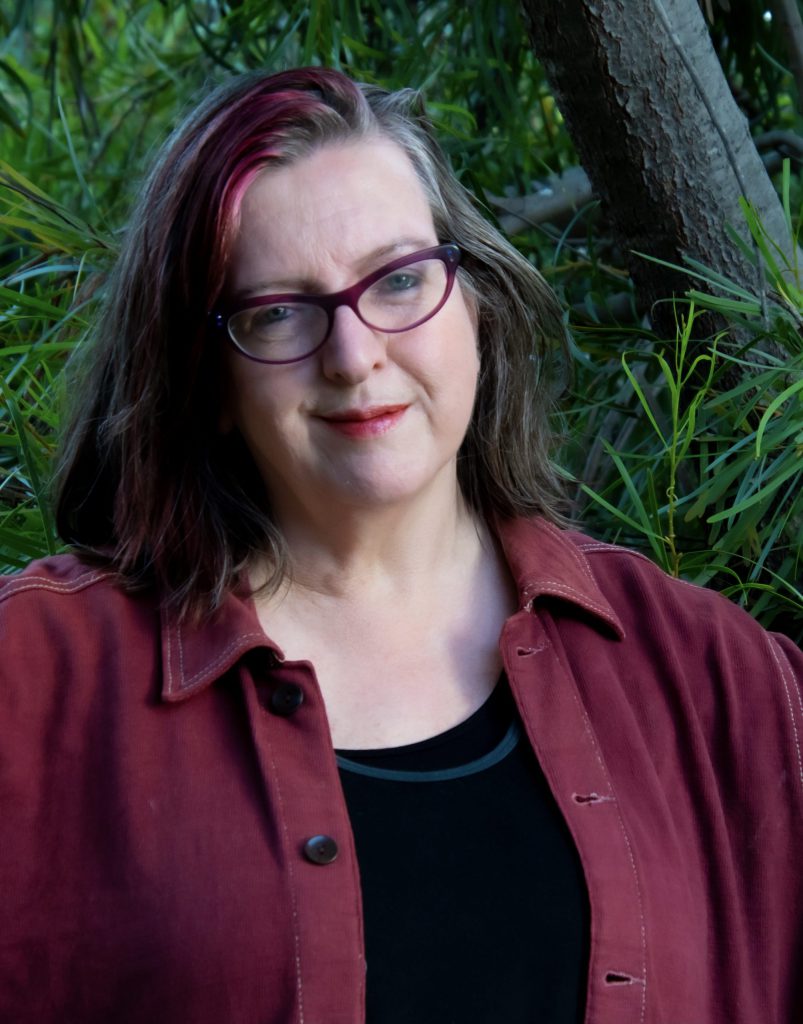 Sarah St Vincent Welch - A smiling woman with shoulder length grey hair, pink at the front, with glasses, wearing a maroon jacket, her arms open, with a leafy backdrop.